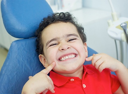 Child in red shirt pointing to smile in treatment chair