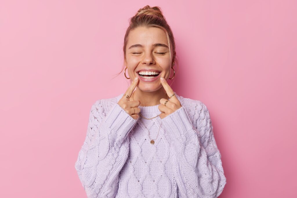 Woman closing eyes pointing to smile with two fingers with pink background